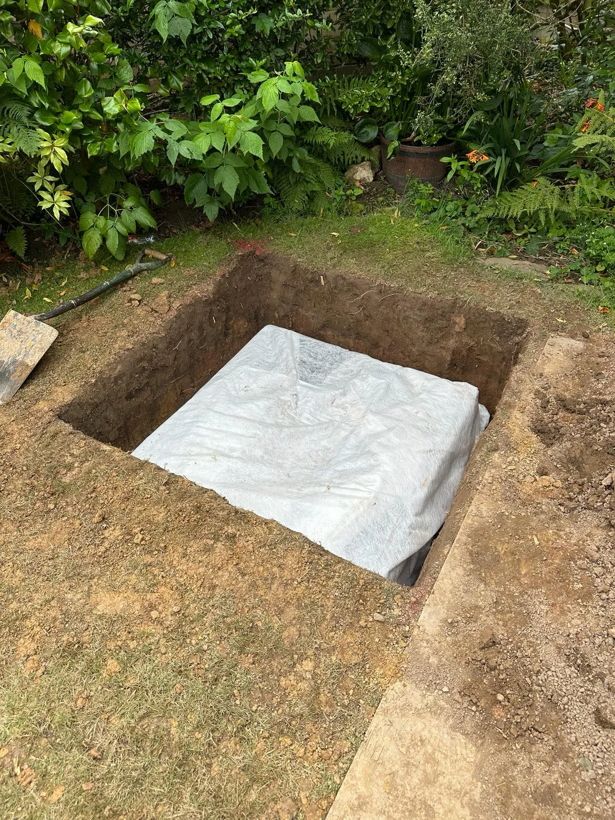 A large pit dug in a garden area containing a white-covered crate structure, possibly a soakaway or attenuation tank, with a shovel leaning nearby.